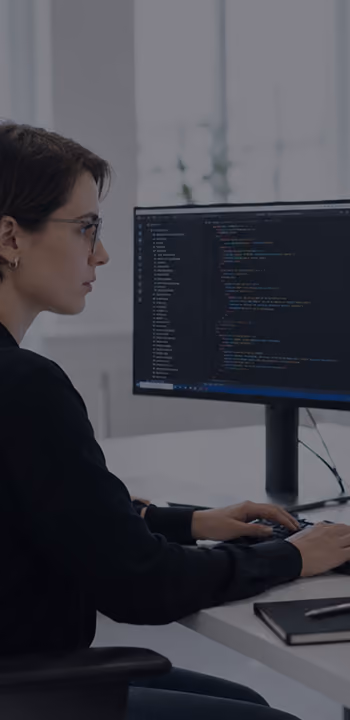 A person with short dark hair and glasses is sitting at a desk working on coding displayed on a large computer monitor in a bright office environment.