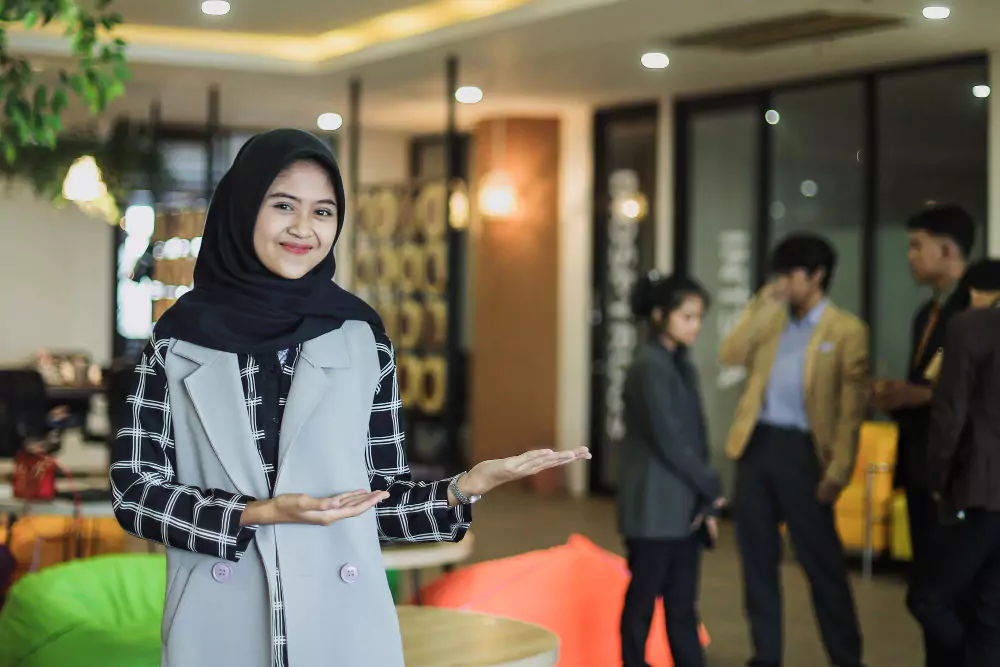 Potret wanita muslimah muda dalam sut perniagaan tersenyum dan menyambut dengan latar belakang pejabat