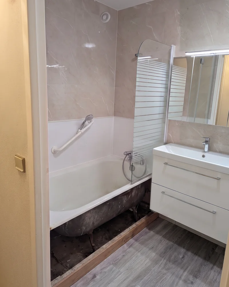 Avant&nbsp;: la&nbsp;baignoire vue depuis la&nbsp;porte, avec un&nbsp;pare-baignoire vitré et&nbsp;une barre d'appui au&nbsp;mur