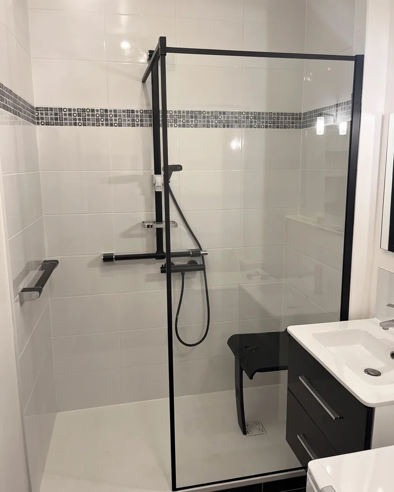 Après&nbsp;: une douche à&nbsp;paroi vitrée cerclée de&nbsp;noir, avec une colonne de&nbsp;douche, des barres d'appui et&nbsp;un&nbsp;siège