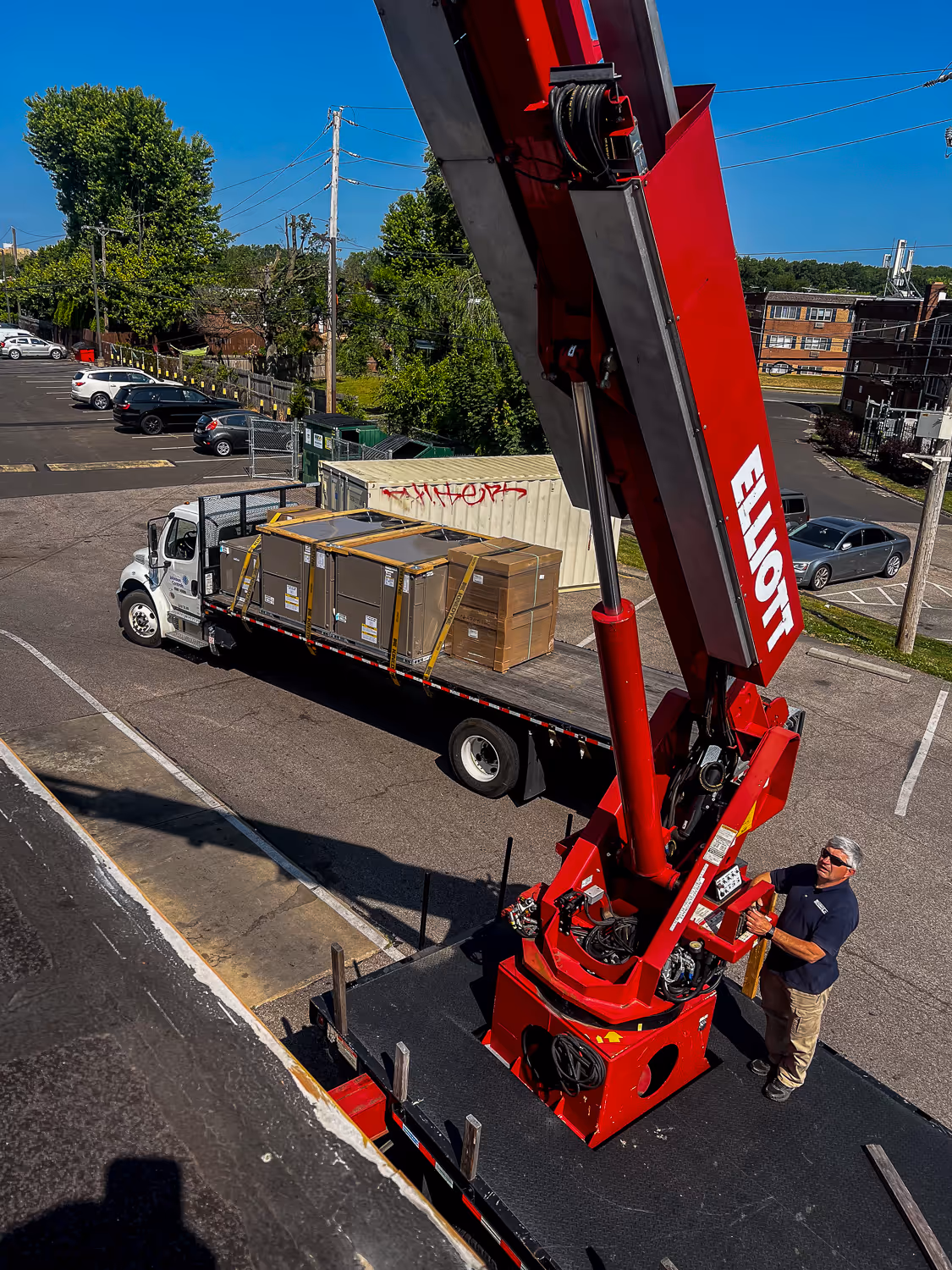 A man operates a red crane to lift a large box from a truck in a parking lot.