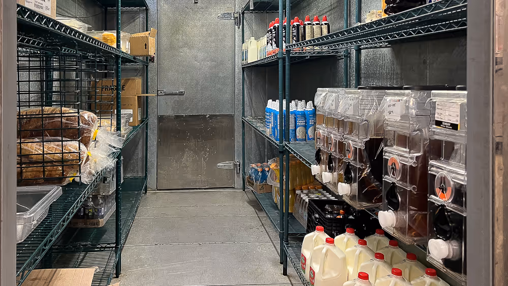 A storage room with metal shelves holding beverages, milk jugs, boxes, and various supplies.