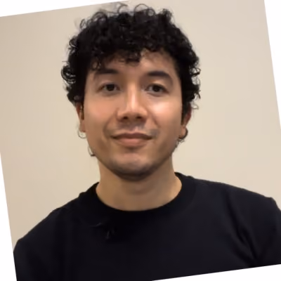 Portrait of a man with curly black hair wearing a black shirt against a beige wall.