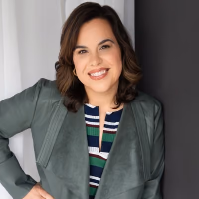 Smiling woman with dark hair wearing a green jacket and striped top against dark and light background.