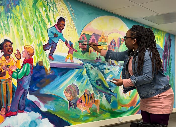 Tia is painting a colorful mural with fish, people, and other wildlife. The mural is vibrant and colorful.