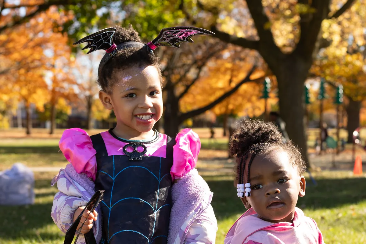 Two young girls in Halloween costumes, one wearing a bat-wing headband, smile under bright fall trees.