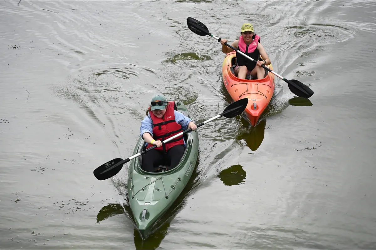 Two paddlers in life jackets kayak toward the camera, one in a green kayak and one in an orange kayak.