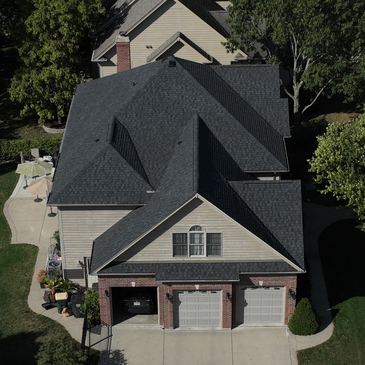 Aerial view of a suburban two-story house with a dark gray shingled roof, beige siding, and a three-car garage, surrounded by trees and lawn.