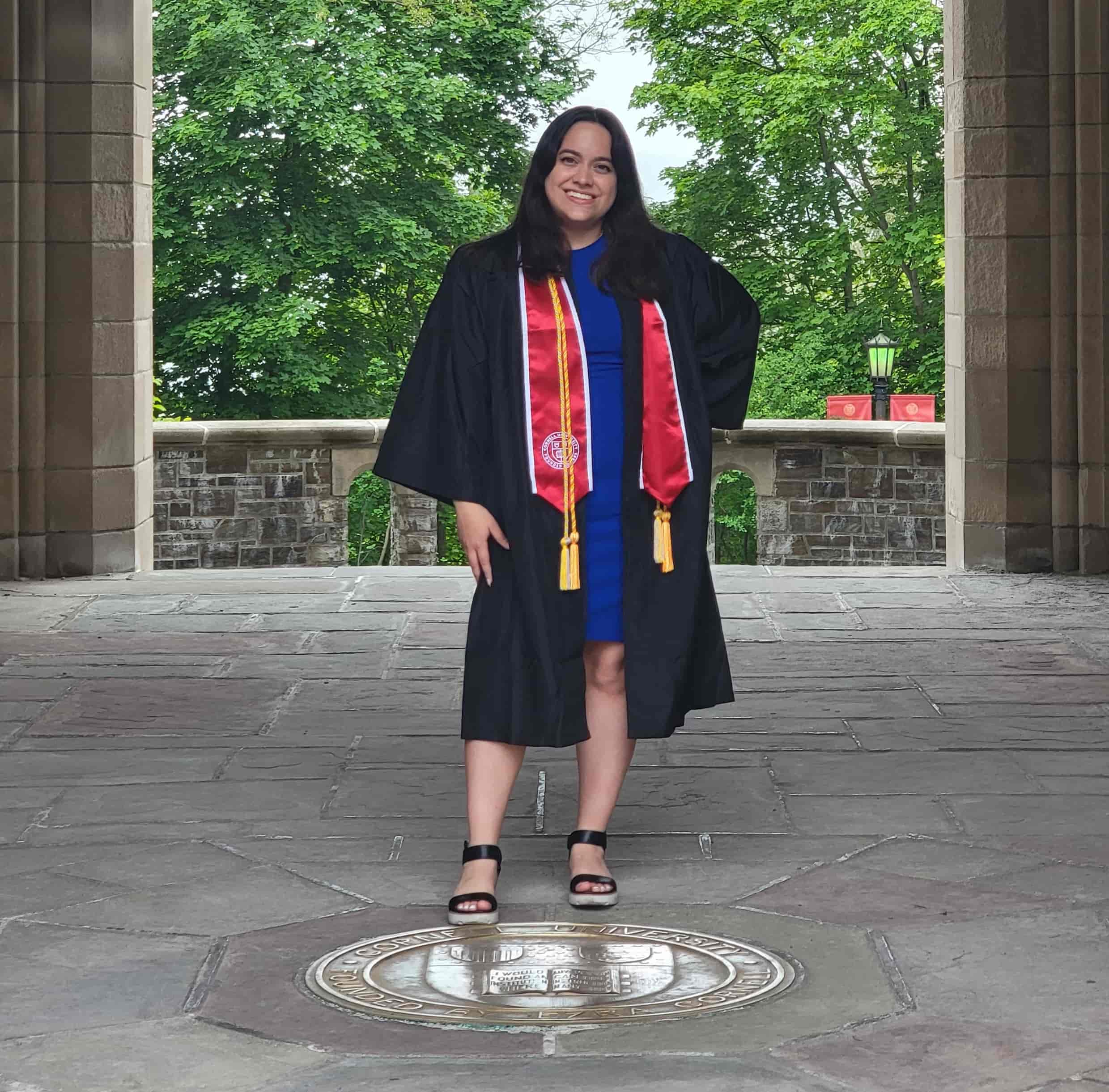 Image of me in front of Cornell logo wearing graduation gown with a Cornell stole

