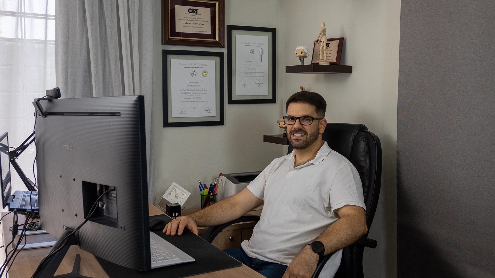 Fotografía de Javier Pereyra Creus, el fundador y principal Motion designer de The Motion Studios. En la imagen, Javier está en su oficina en Montevideo, Uruguay, sonriendo luego de terminar un video explicativo con animación y motion graphics, mirando la cámara sentado en su silla en su escritorio frente a su computadora y rodeado de sus diplomas de Licenciado en Diseño Multimedia y el premio al desempeño académico por la Universidad ORT Uruguay. El personaje principal de The Motion Studios de color azul oscuro y forma de cubo con ojos grandes y blancos y brazos y piernas de fideo o palitos está sobre su escritorio saludando a la cámara.