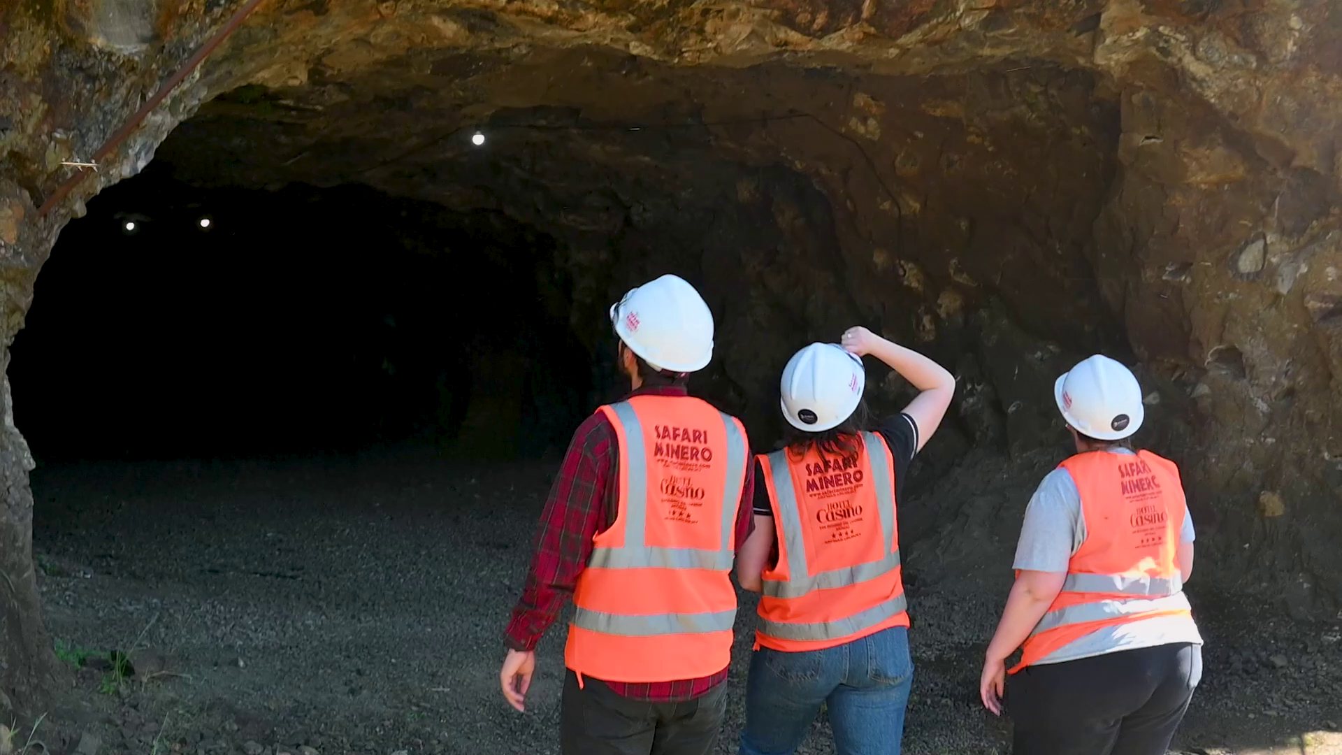 Tres personas con cascos blancos y chalecos naranjas miran hacia la entrada de una cueva oscura.