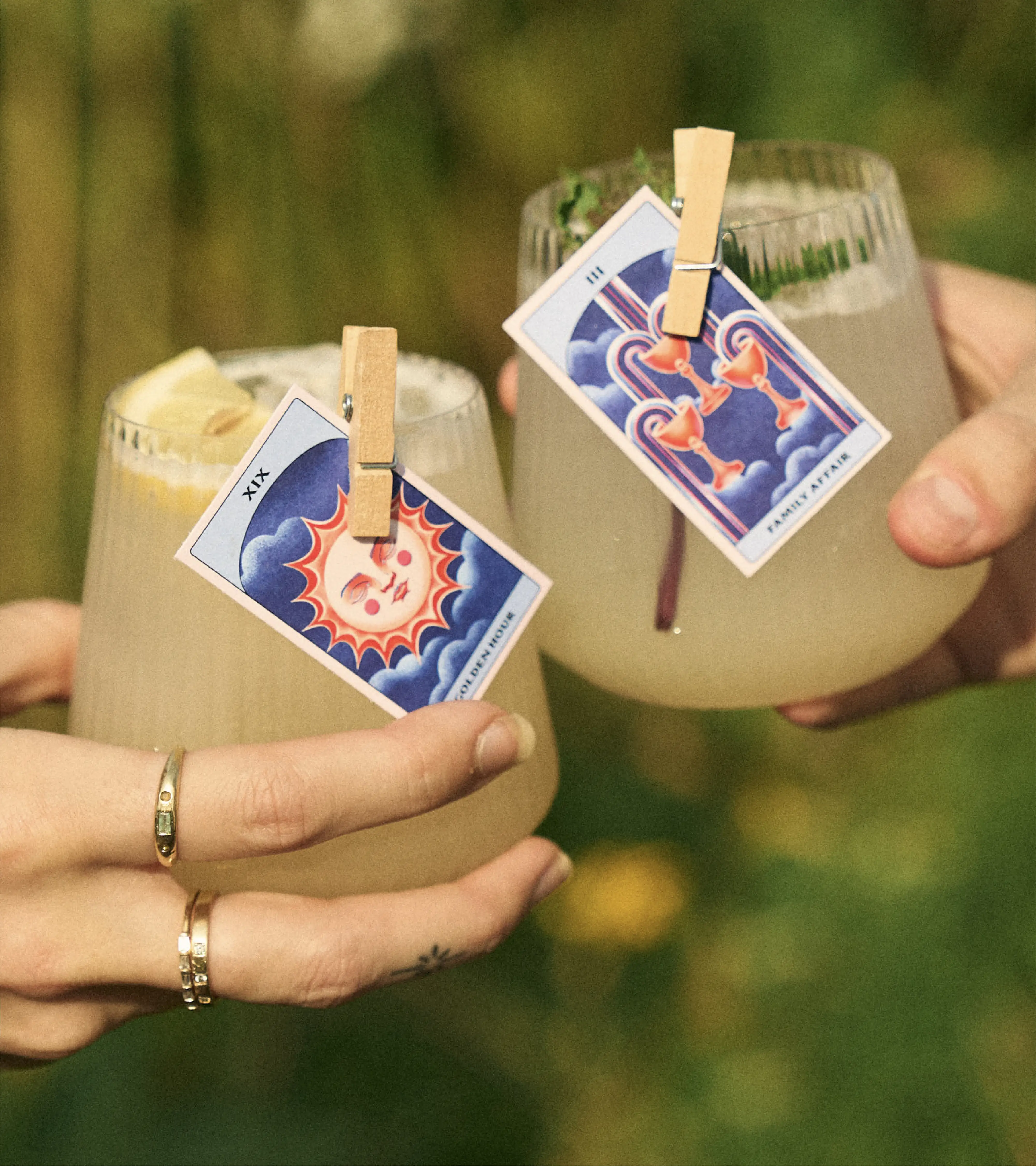 Two yellow cocktails cheersing on a green, blurry background. On each of the cocktails in a tarot card clipped to the trim as garnish. On one is The Sun and the other, the Three of Cups