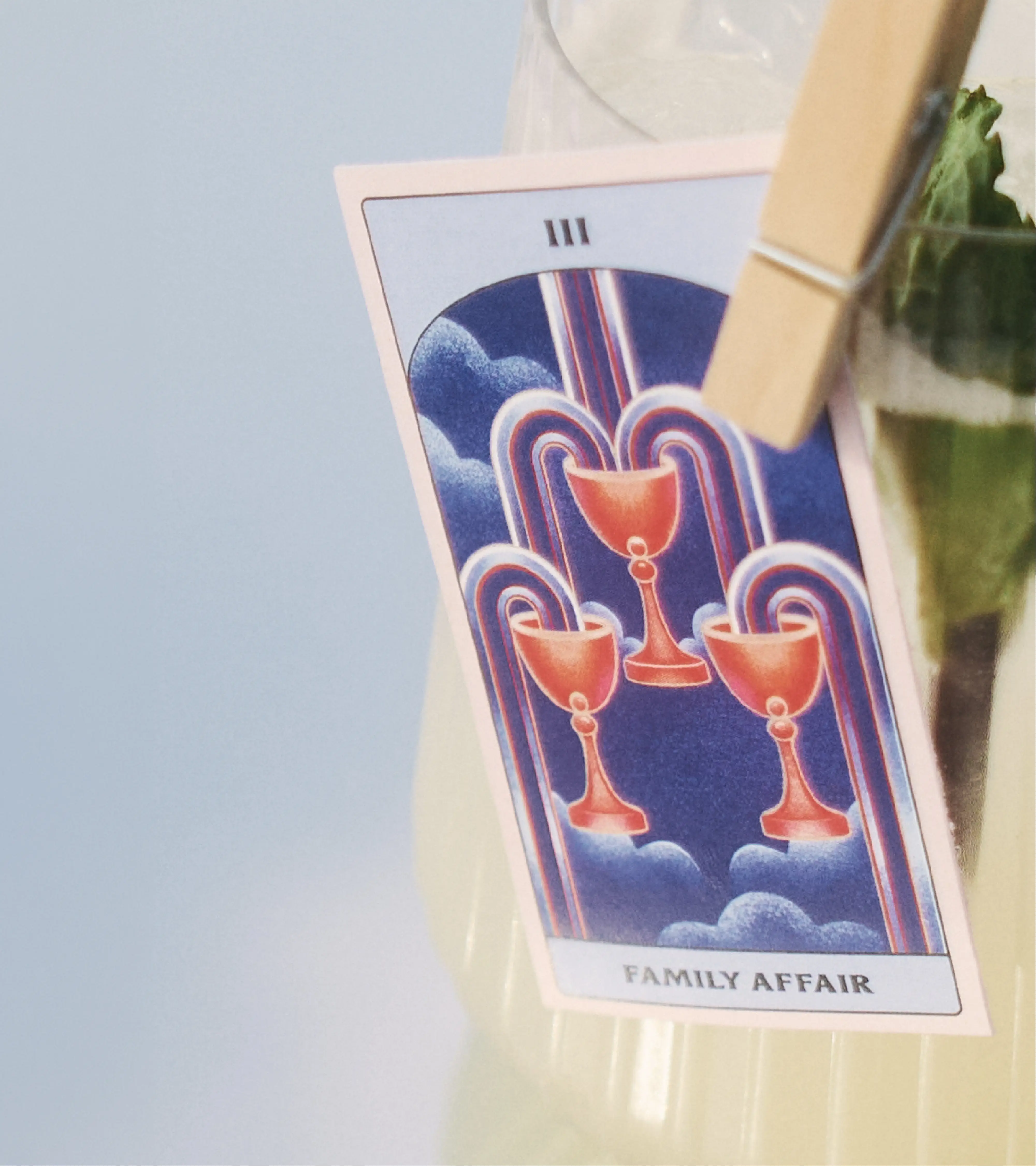 Extreme close up of the tarot card garnish clipped to the rim of the glass. In it you can see the illustrations that coincide with the candle design - specifically the three of cups. It is on an ethereal, blurry blue background