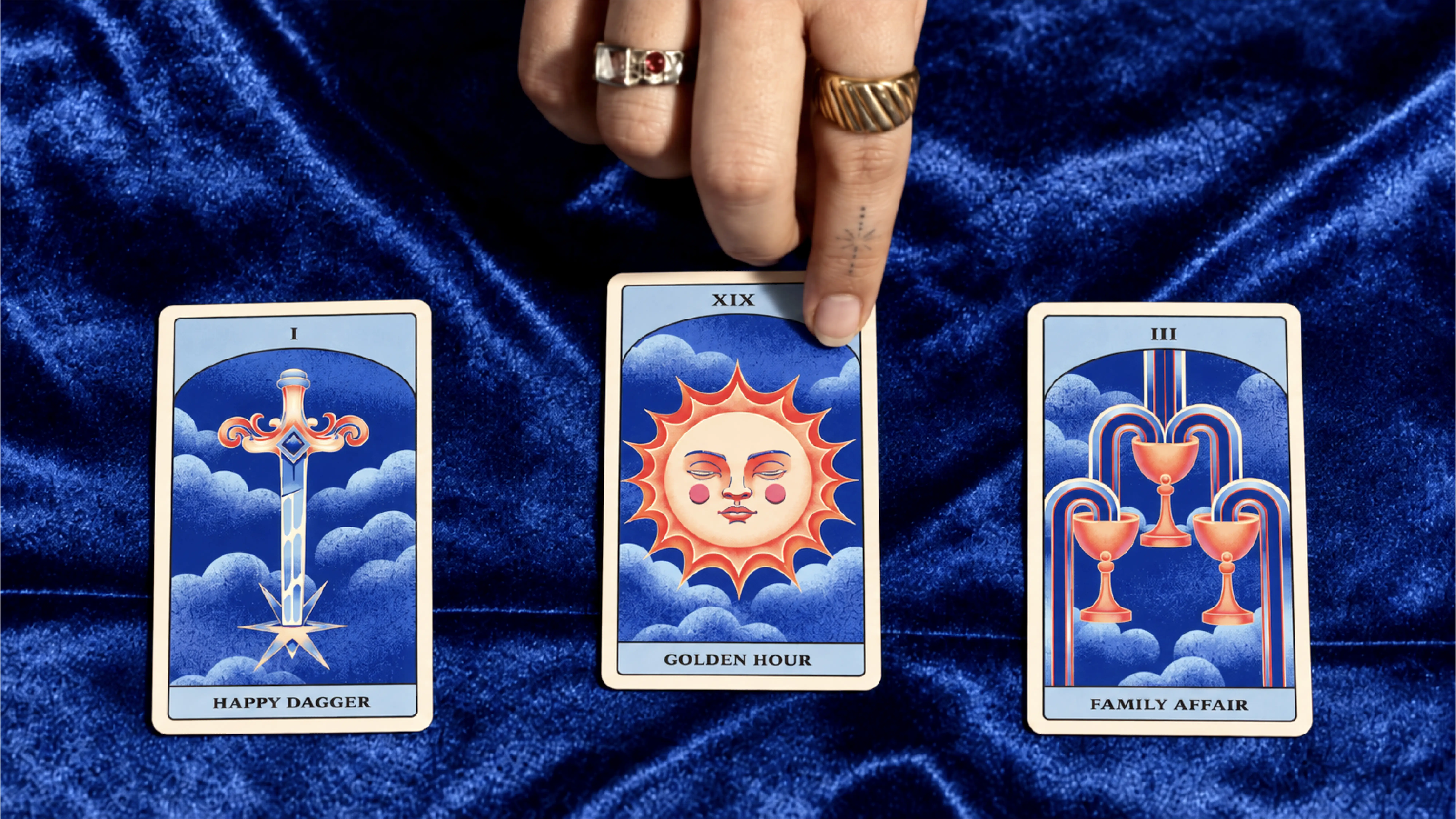 Three tarot cards shown on a crushed blue velvet background. The three cards are the Ace of Swords, The Sun, and the Three of Cups. A hand is reaching in from the top of the frame and pulling up on the Sun card.