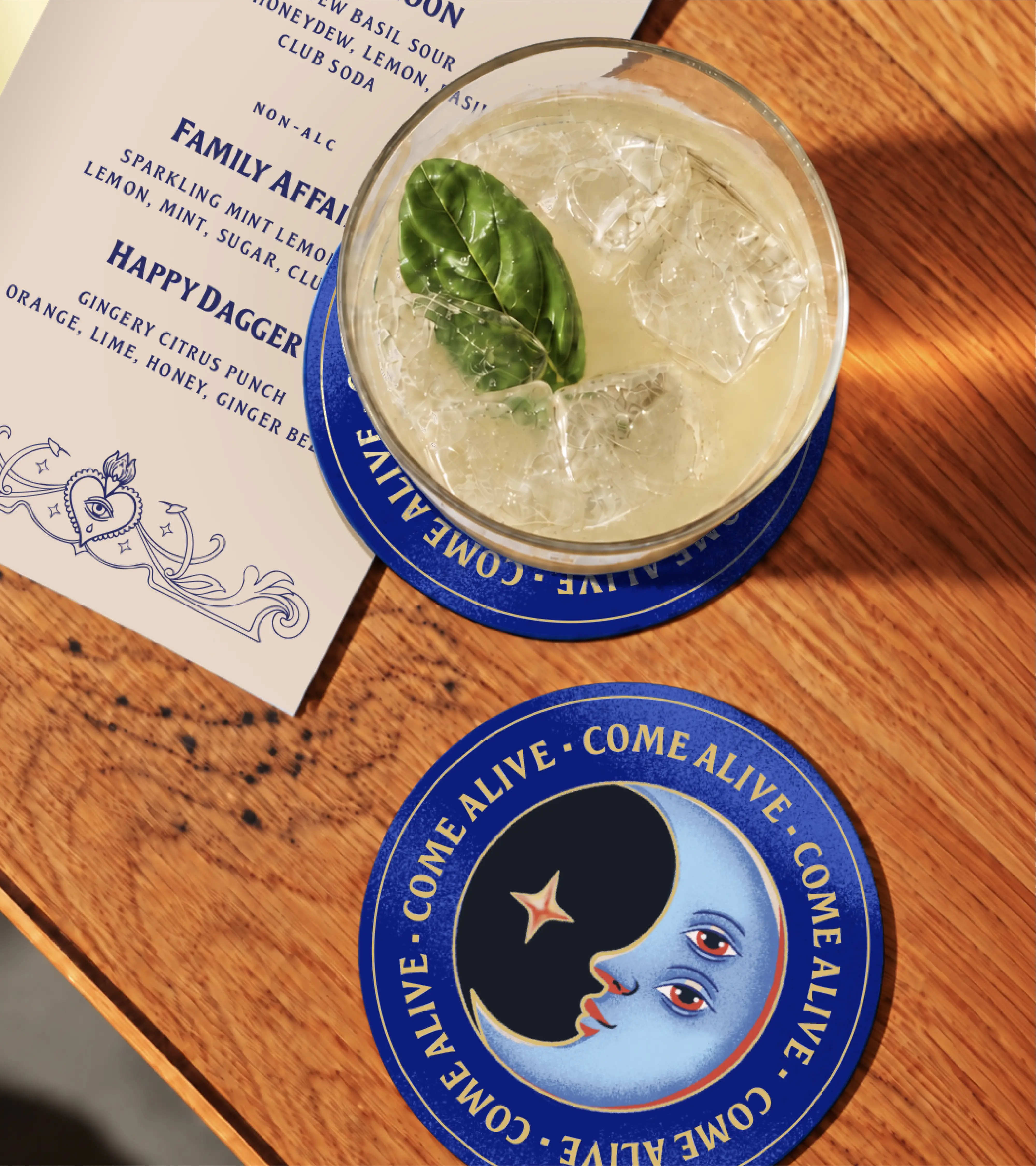 Top-own photo of a cocktail garnished with bail on a wooden surface next to a blue branded coaster and a menu. The coaster features the moon illustration with the words "come alive" repeated around the perimeter.