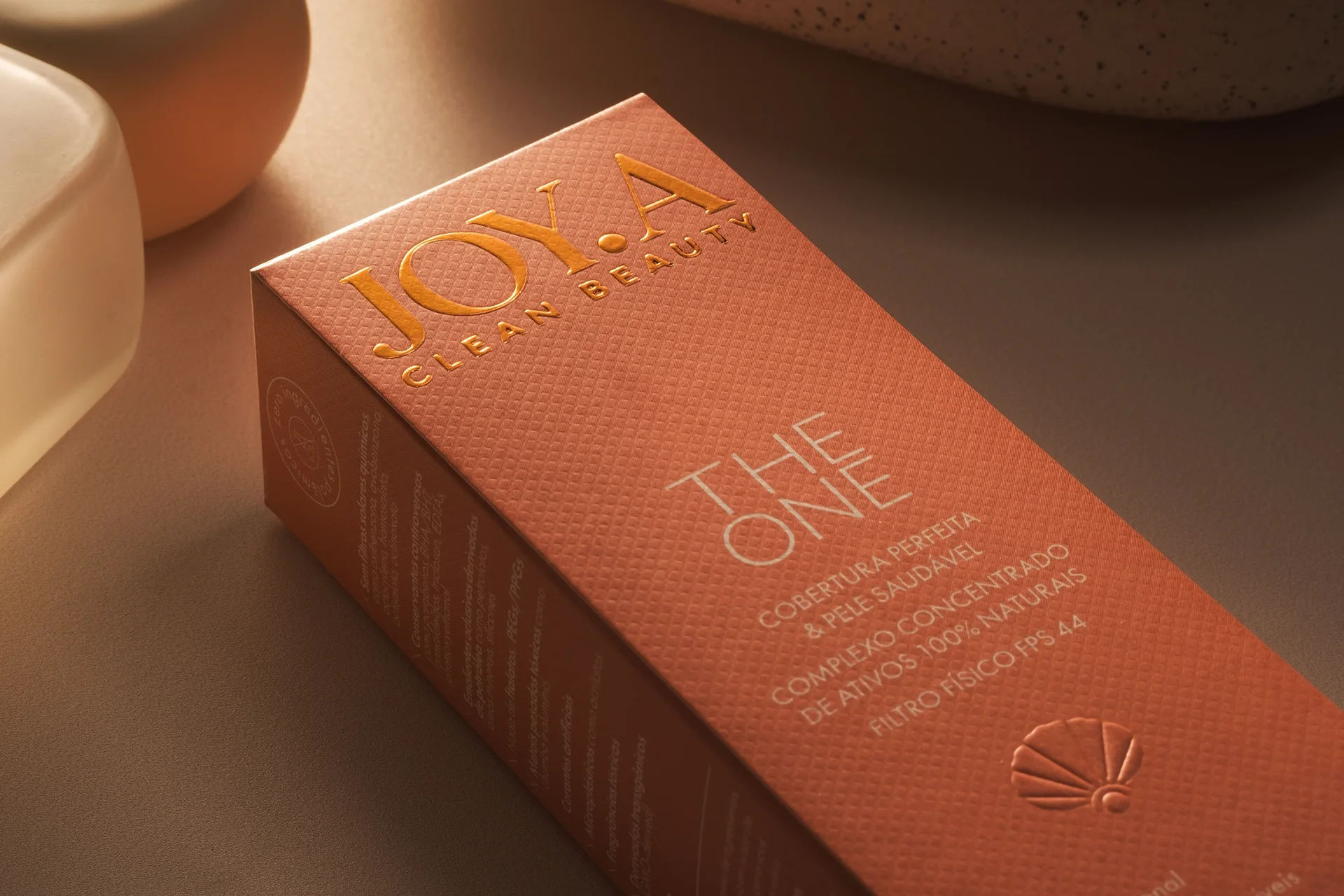 Design de caixa de cosmético "THE ONE", em tom pêssego com letras douradas. Levemente inclinada, revela textura e logo com concha. Luz suave destaca a embalagem sobre superfície neutra.