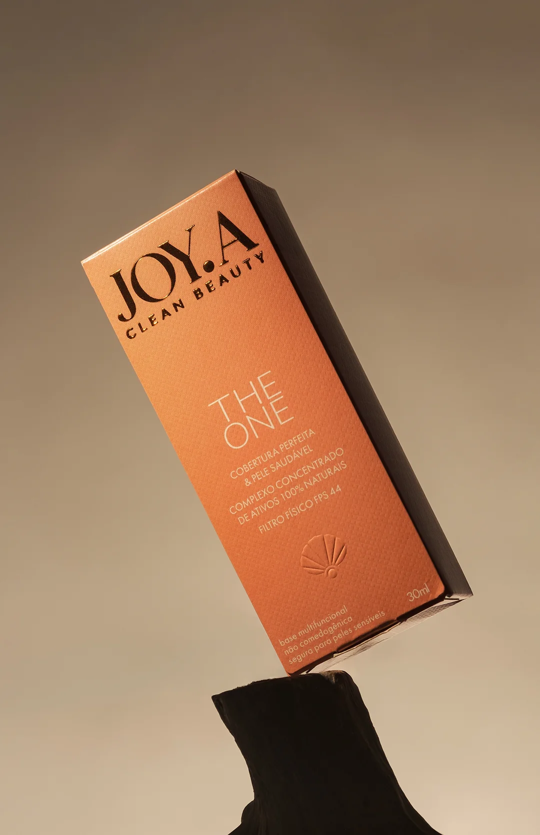 Design de caixa de cosmético "THE ONE", em tom pêssego com letras douradas. Levemente inclinada, revela textura e logo com concha. Luz suave destaca a embalagem sobre superfície neutra.