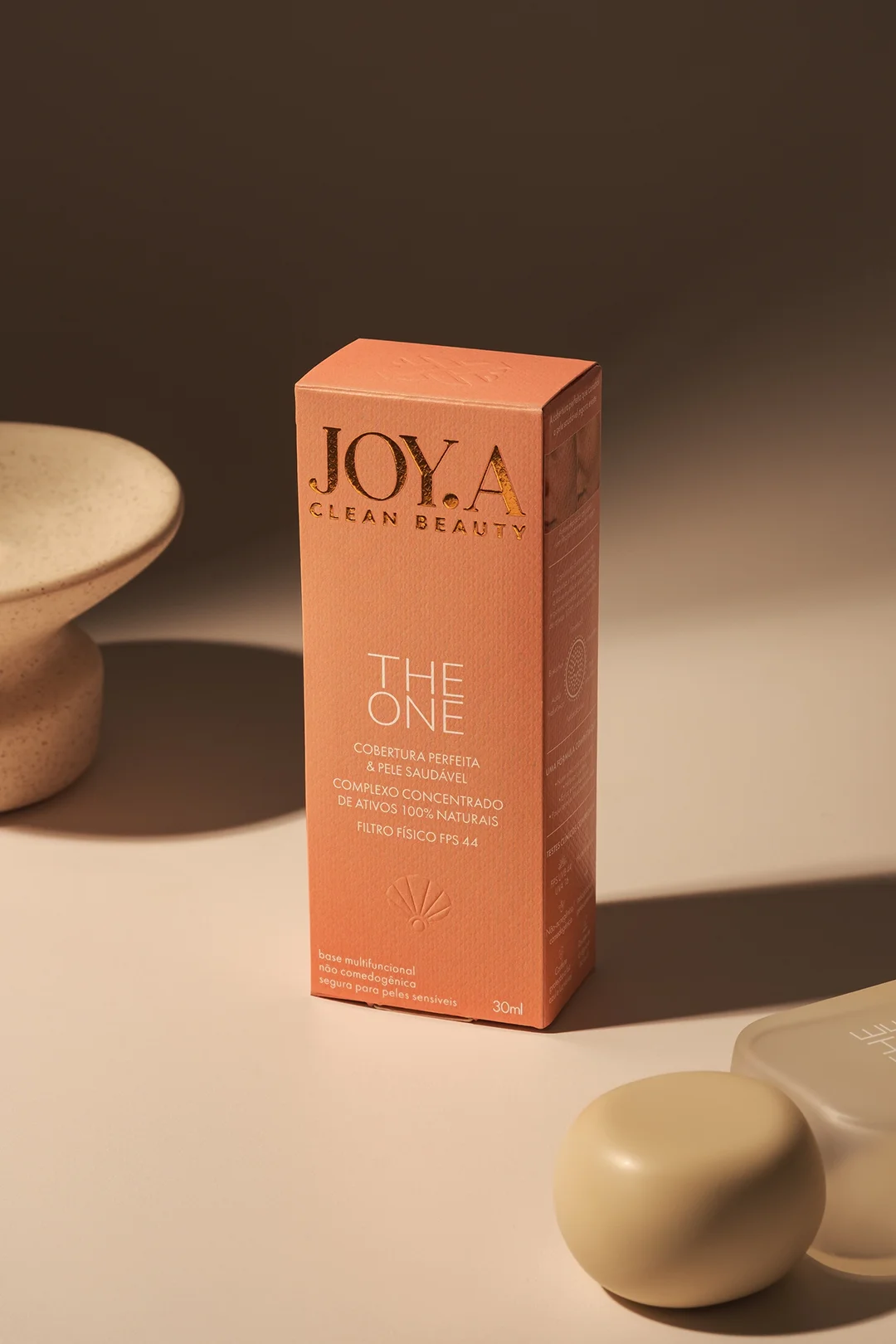 Design de caixa de cosmético "THE ONE", em tom pêssego com letras douradas. Levemente inclinada, revela textura e logo com concha. Luz suave destaca a embalagem sobre superfície neutra.
