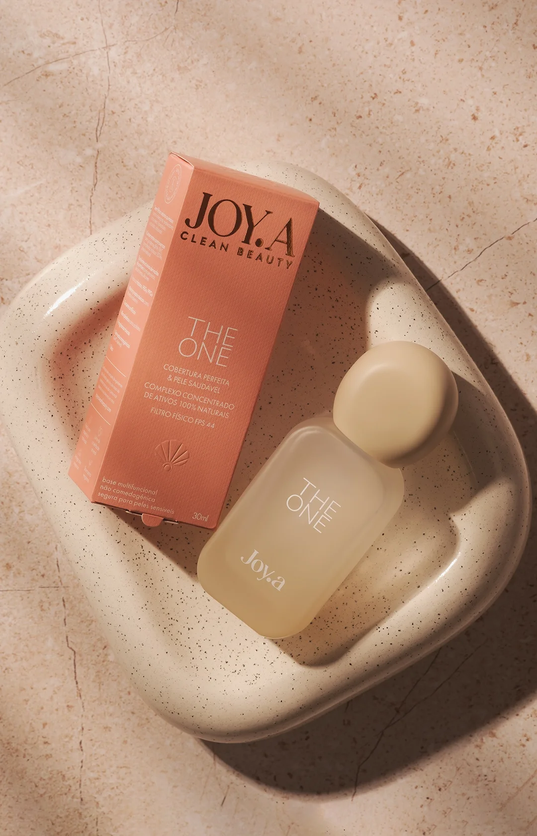 Um frasco cosmético bege claro com o rótulo "JOY.A THE ONE" ao lado de uma embalagem laranja, sobre um prato raso e salpicado. A composição, em mármore claro, destaca a estética clean beauty.