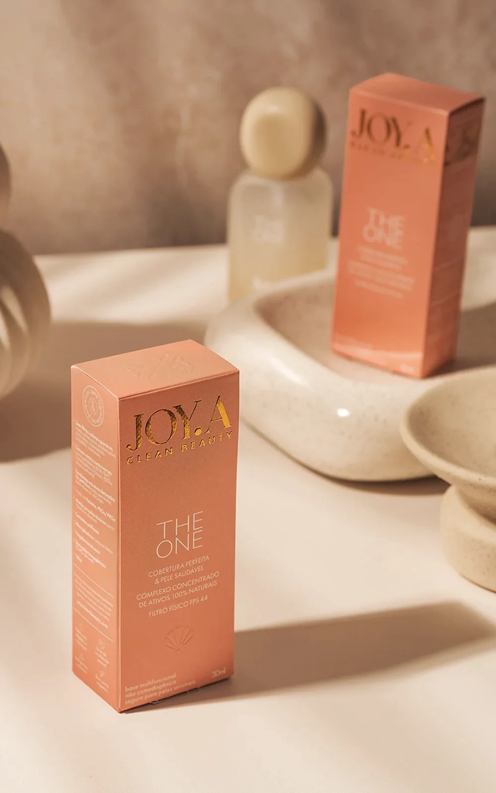 design dos produtos JOYA, com duas caixas "THE ONE" e um frasco transparente. Tons neutros com toque laranja sobre fundo texturizado, destacando o design da embalagem.