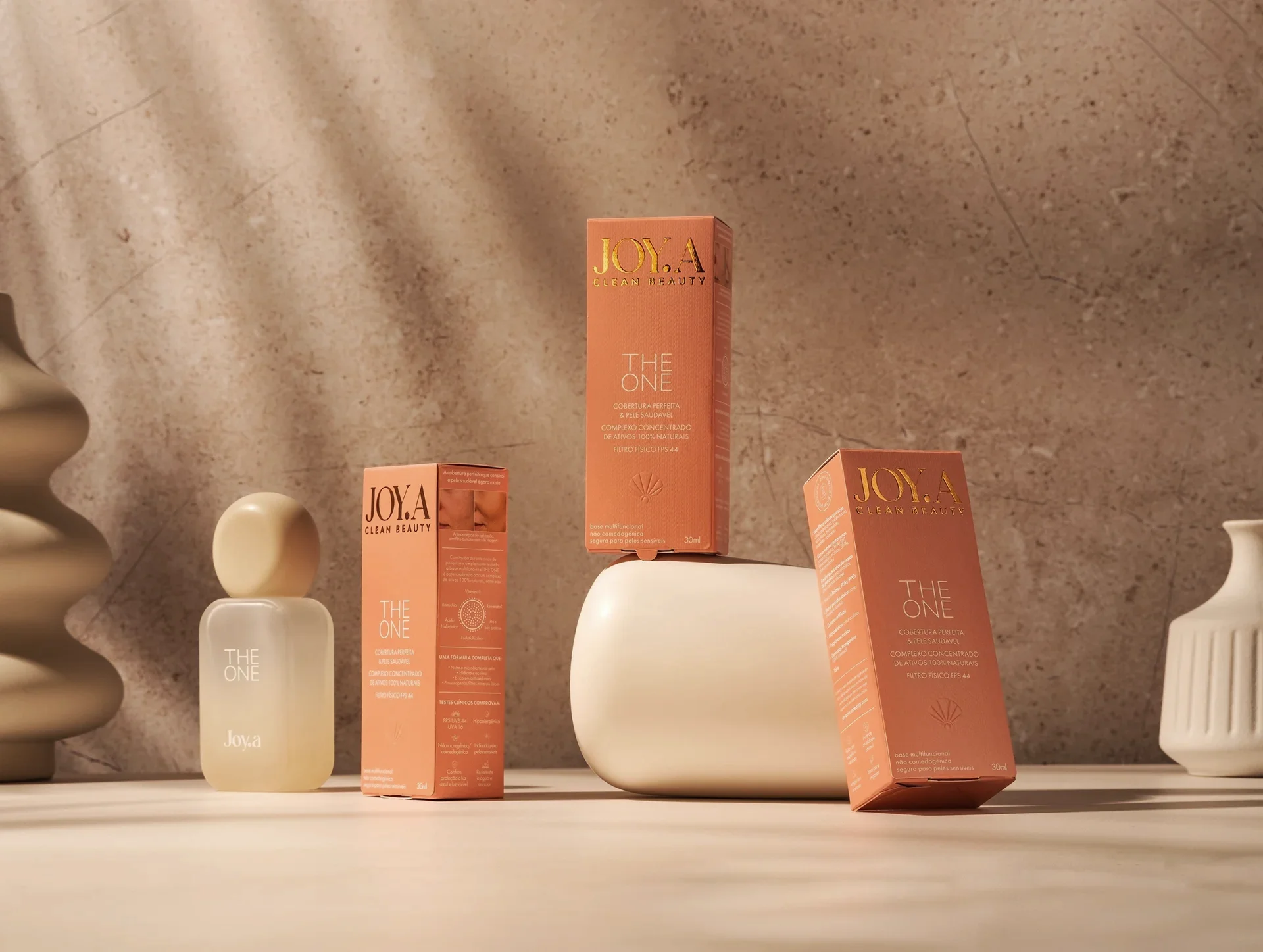 Design dos produtos JOYA, com duas caixas "THE ONE" e um frasco transparente. Tons neutros com toque laranja sobre fundo texturizado, destacando o design da embalagem.