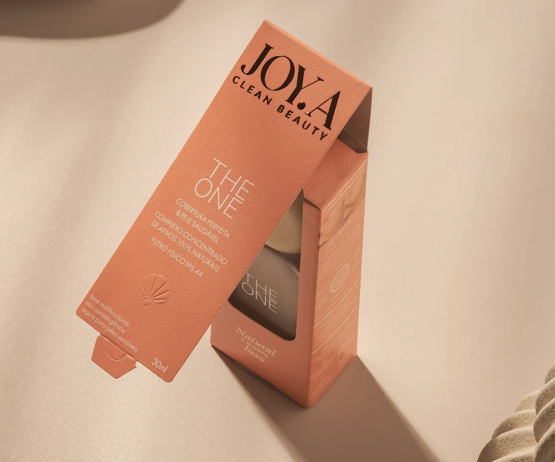 Design de caixa de cosmético "THE ONE", em tom pêssego com letras douradas. Levemente inclinada, revela textura e logo com concha. Luz suave destaca a embalagem sobre superfície neutra.