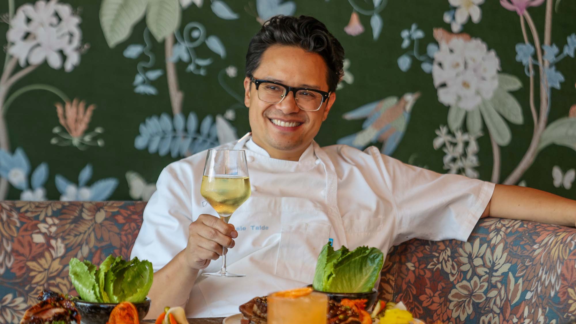 Smiling chef in white uniform holding a glass of white wine, seated at a table with colorful dishes and a floral wallpaper background.