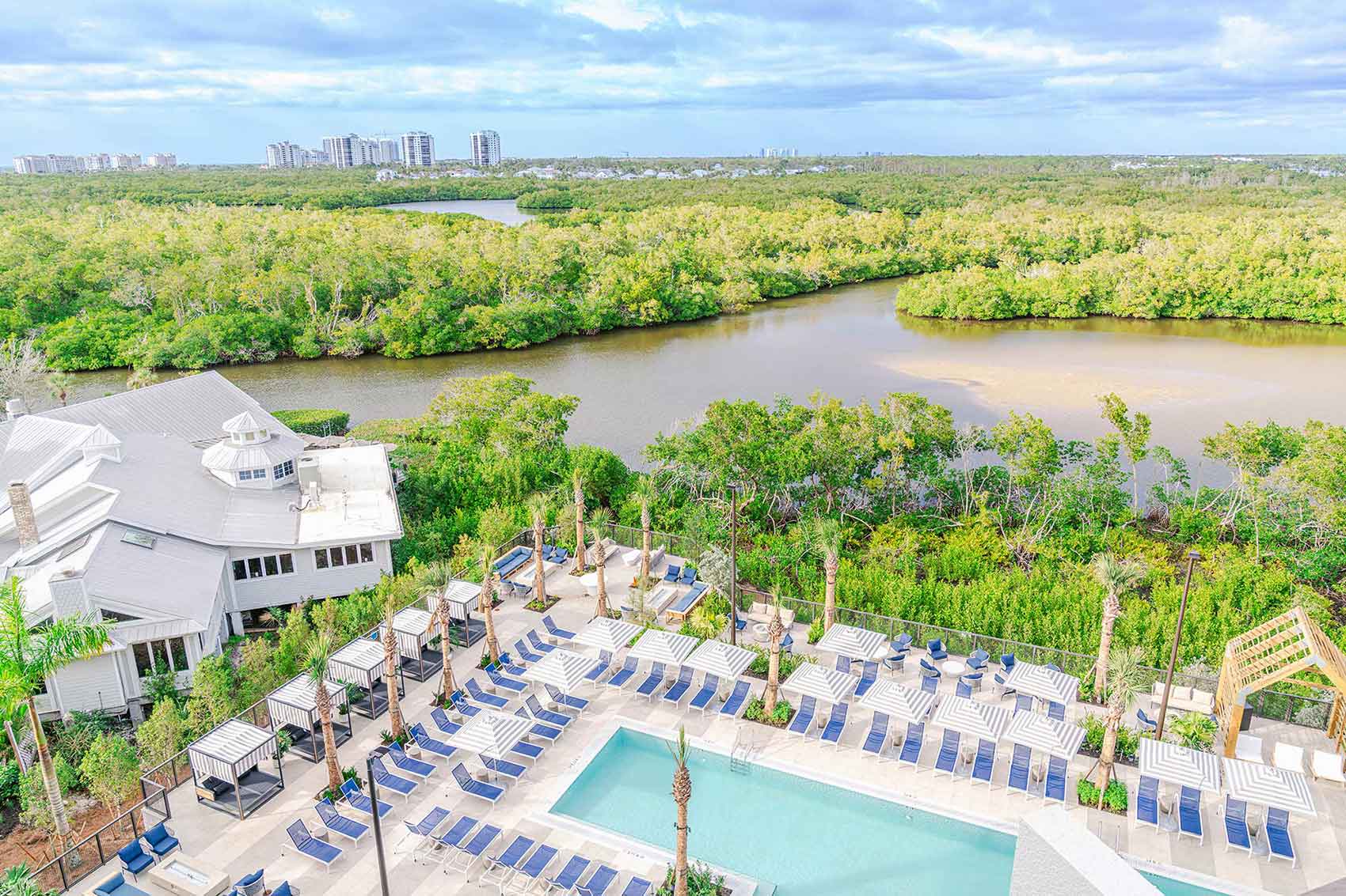 Aerial view of a resort pool area with rows of blue lounge chairs and striped umbrellas, surrounded by lush greenery and a river, with a skyline of high-rise buildings in the distance.