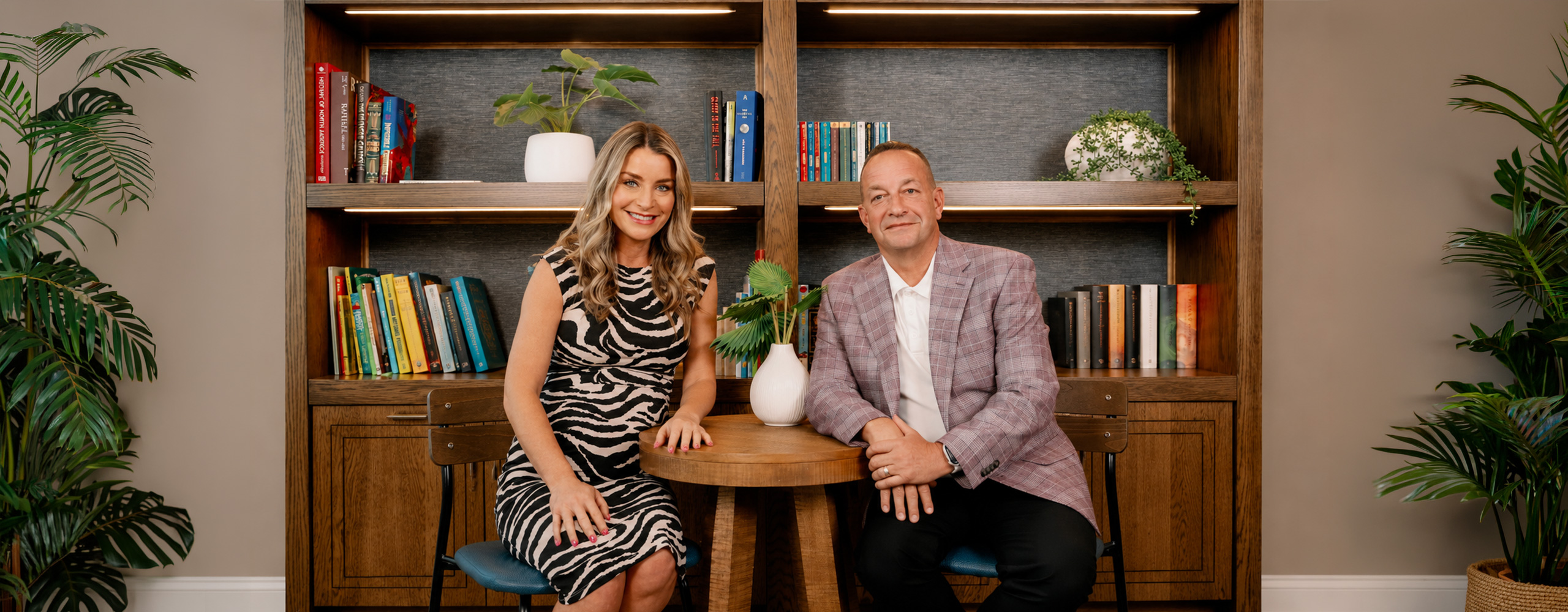 Two people seated at a small round table in front of a styled bookshelf backdrop, smiling toward the camera in a staged interview or office setting.