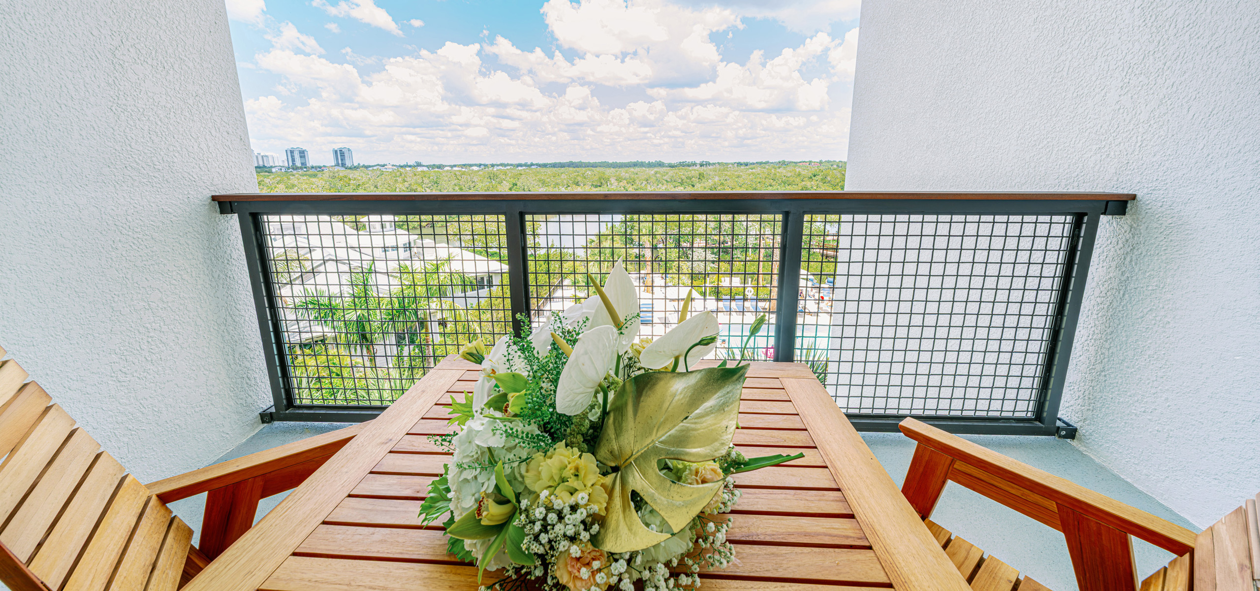 Private hotel balcony with a wooden table and chairs overlooking a resort pool area, greenery, and distant skyline under a bright blue sky. Modern hotel guest room with a large bed, wood-look tile flooring, lounge seating, ceiling fan, and floor-to-ceiling curtains covering a balcony door. Spacious hotel room with two large beds, wood furniture, a mounted TV, patterned tile flooring, ceiling fan, desk area, and sheer curtains over a balcony window. Balcony seating area with a wooden table and chairs, floral centerpiece, mesh railing, and expansive tropical landscape view under a bright blue sky with clouds.