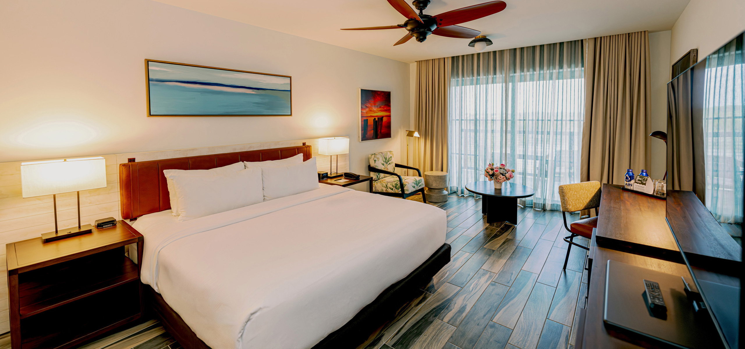 Spacious hotel room with two large beds, wood furniture, a mounted TV, patterned tile flooring, ceiling fan, desk area, and sheer curtains over a balcony window.