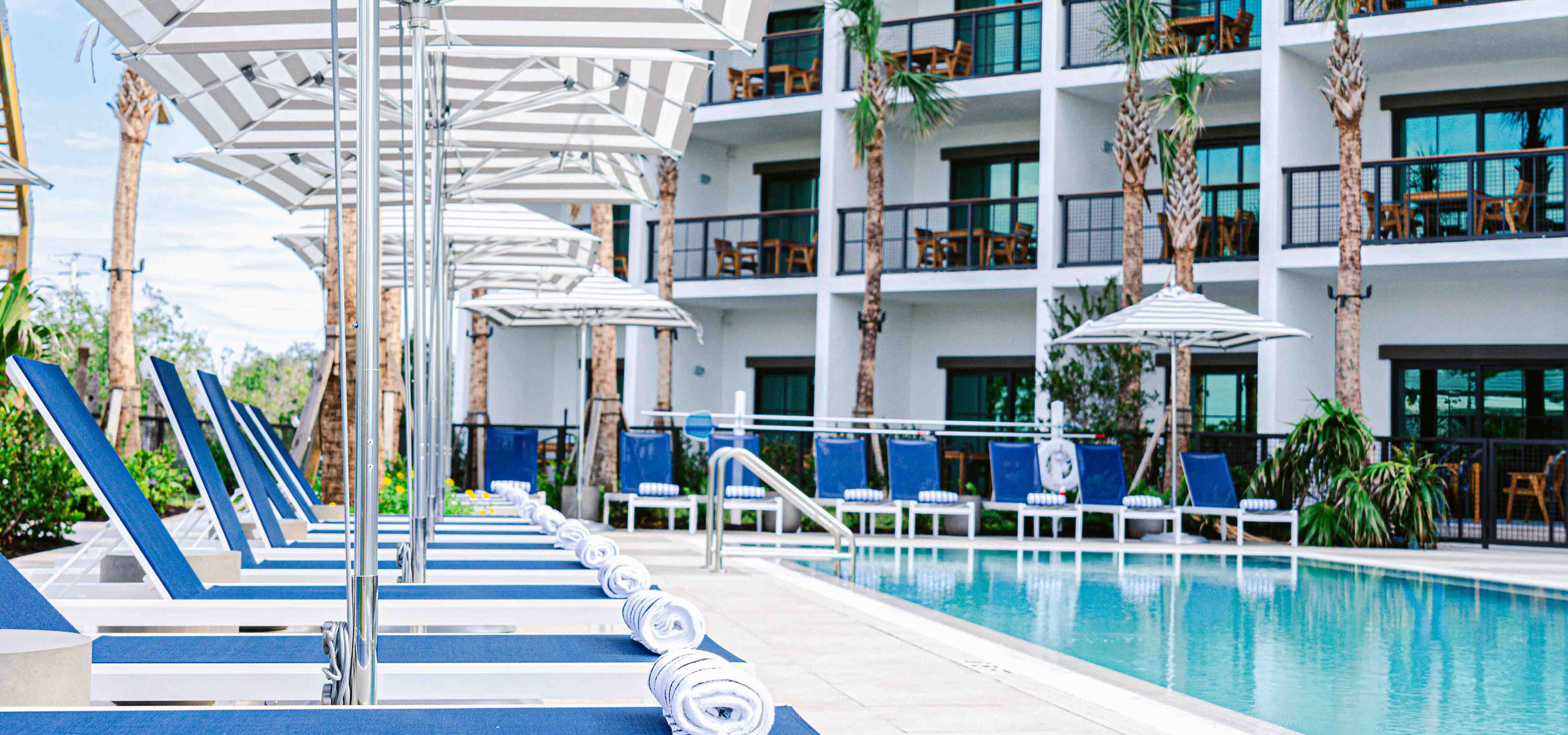 Blue lounge chairs with rolled towels line a resort swimming pool beneath striped umbrellas. A multi-story hotel building rises behind the pool deck.