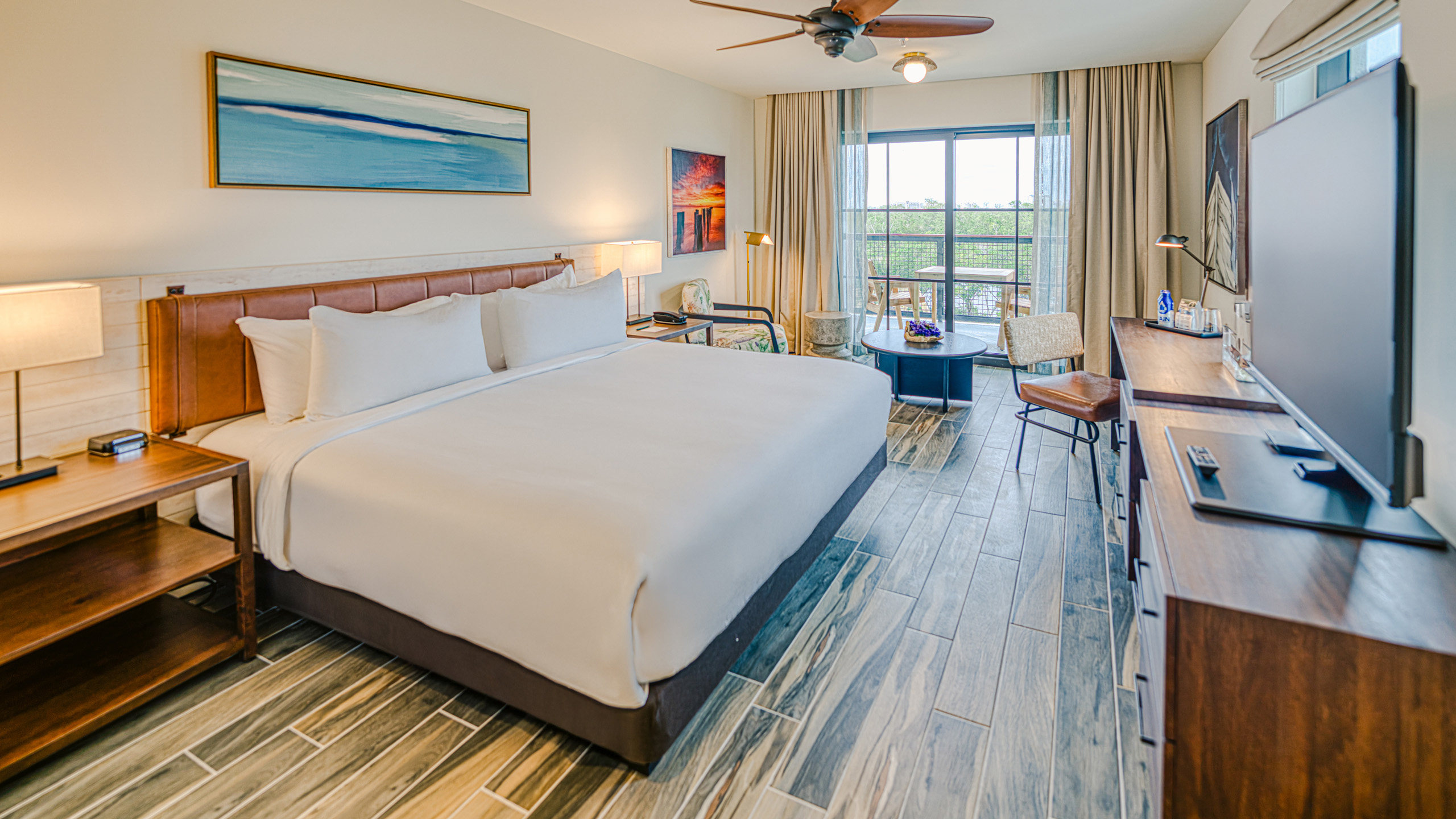 A hotel guest room features a king bed, bedside tables, a desk, seating area, and a furnished balcony. Large windows bring natural light into the modern space.