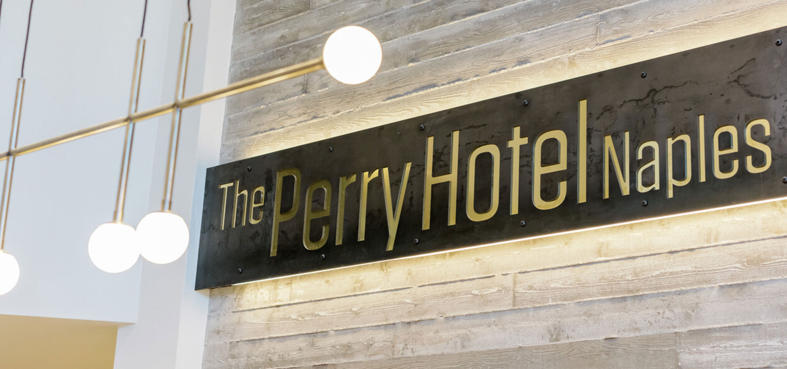 The Perry Hotel Naples sign is mounted on a wood-paneled wall beneath globe pendant lights.