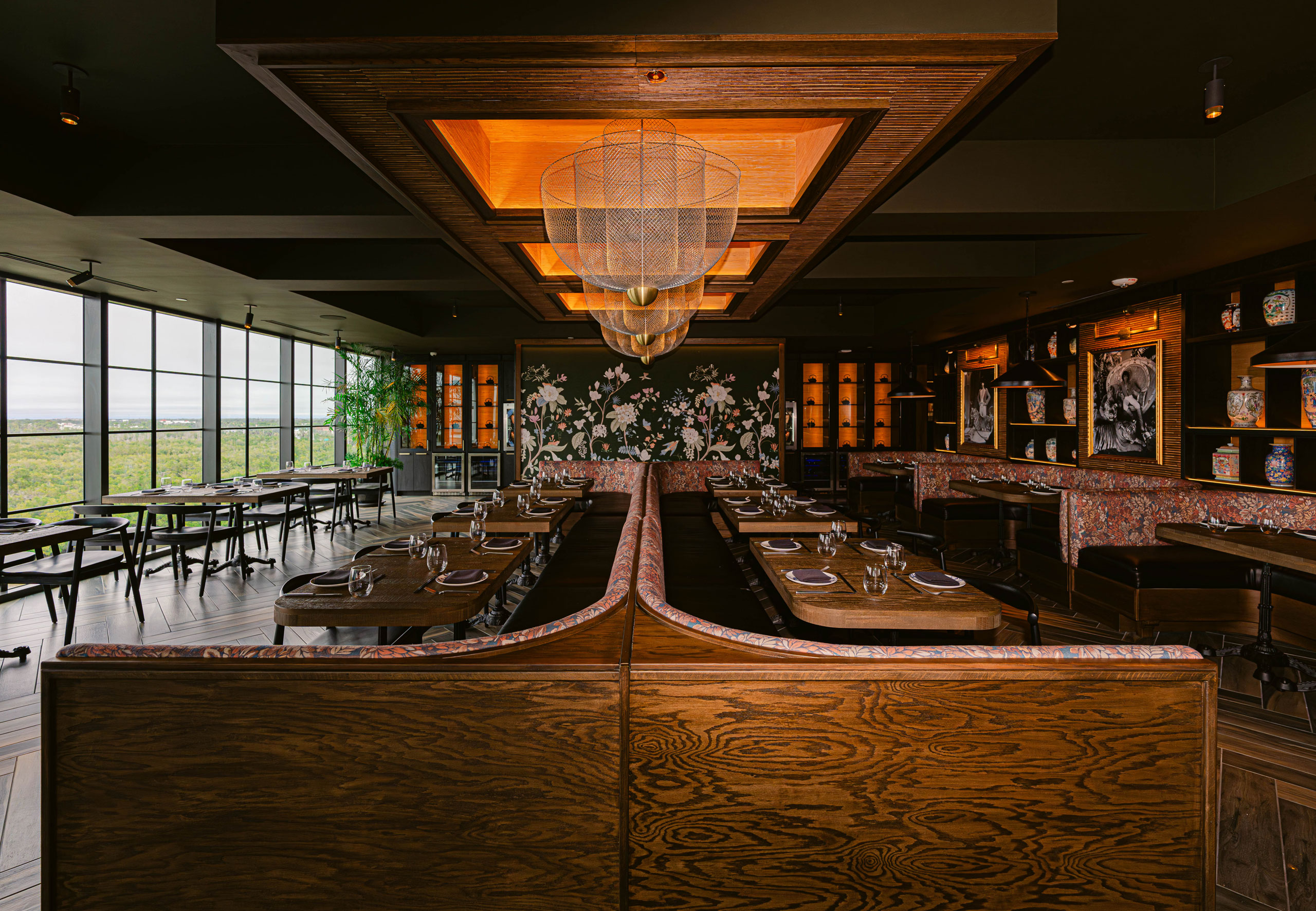 Restaurant interior with patterned booths, wooden tables, pendant lighting, and floor-to-ceiling windows overlooking greenery.