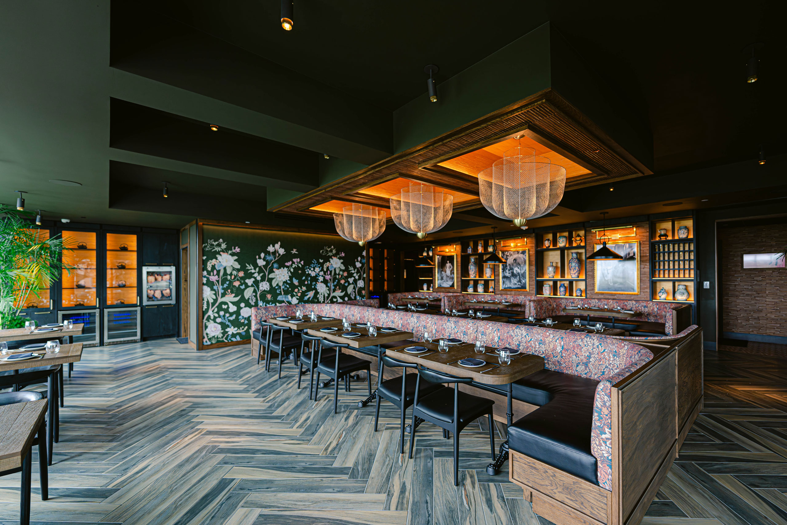 Restaurant interior with floral booths, wooden tables, decorative lighting, and illuminated display shelves.