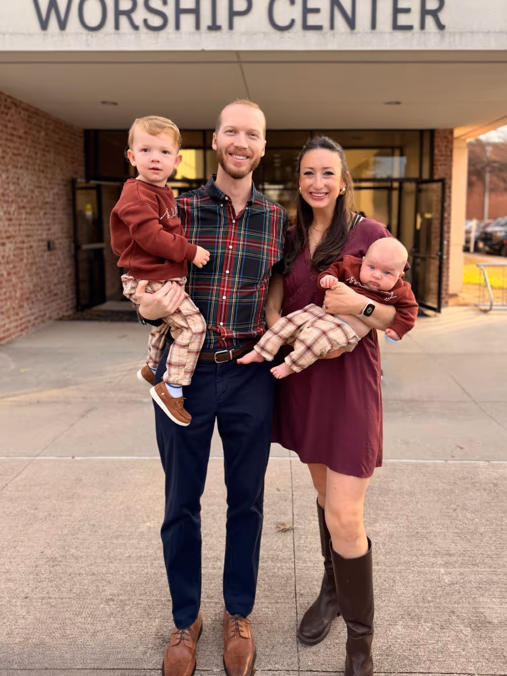 A smiling family of four standing outside a worship center, with a man holding a toddler and a woman holding a baby.