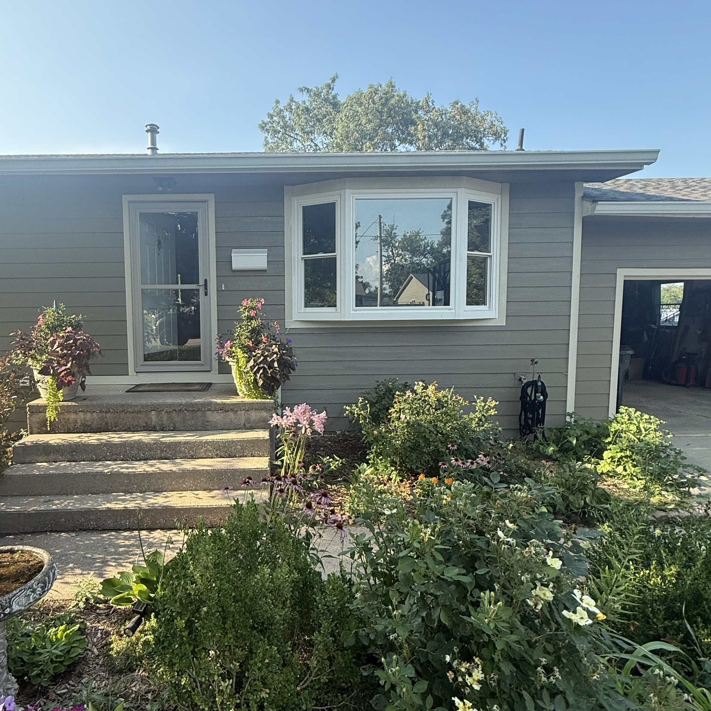 Front view of a single-story house with gray siding, a bay window, concrete steps, and a garden with assorted plants and flowers in the foreground.