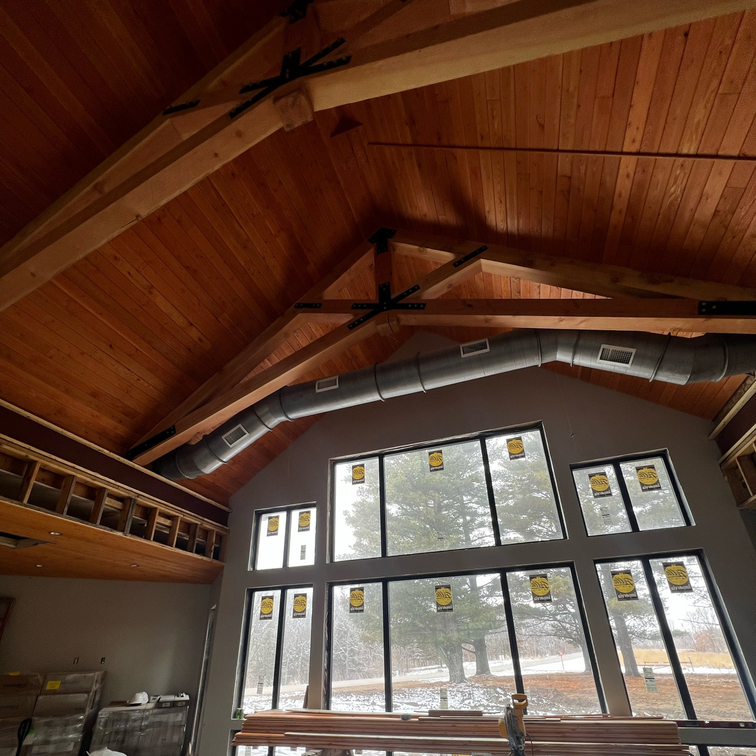 Interior of a building under construction featuring a high wooden ceiling with exposed beams and ductwork, large windows with construction stickers, and construction materials inside.