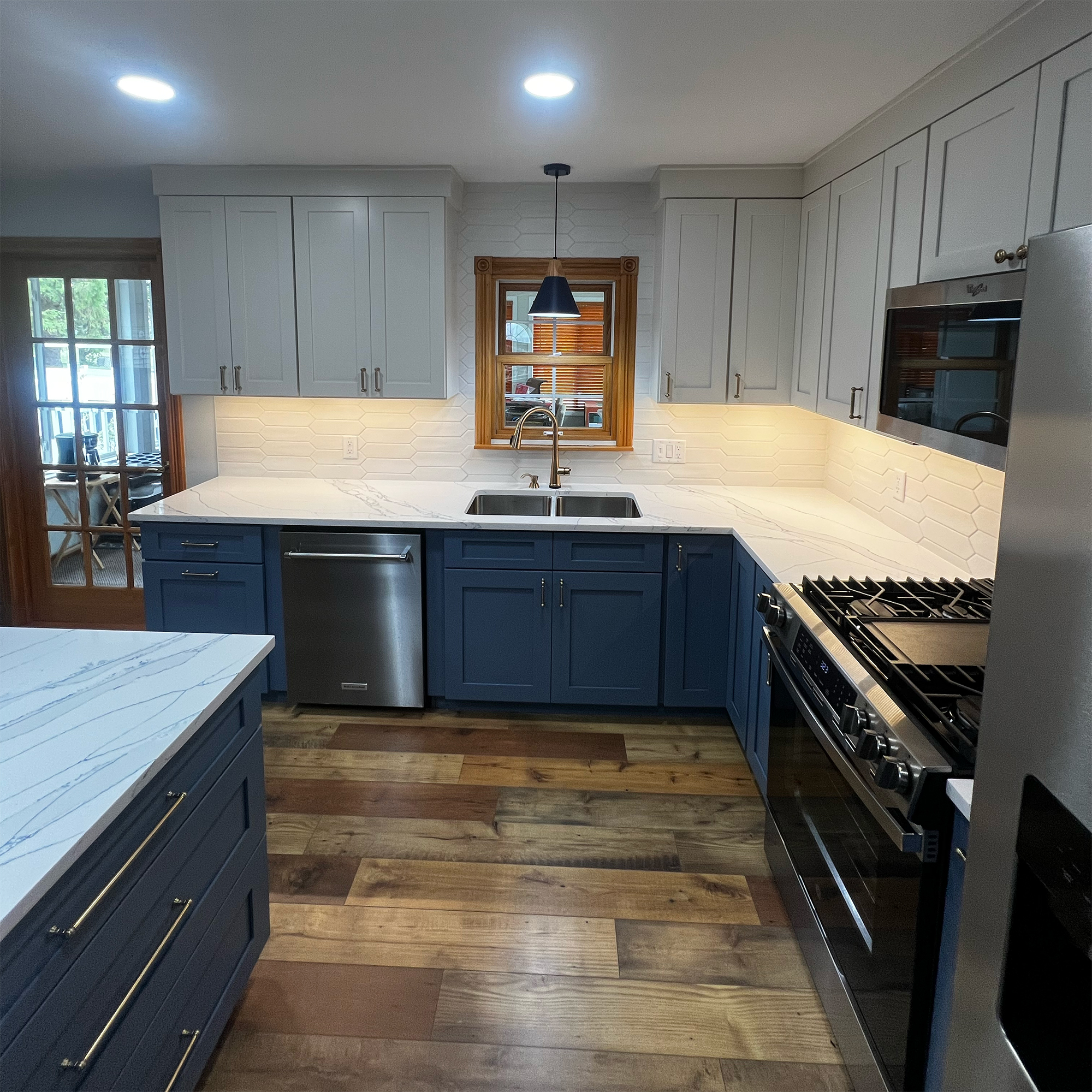 Modern kitchen with blue and white cabinets, white marble countertops, stainless steel appliances, wooden floor, and a window above the sink.
