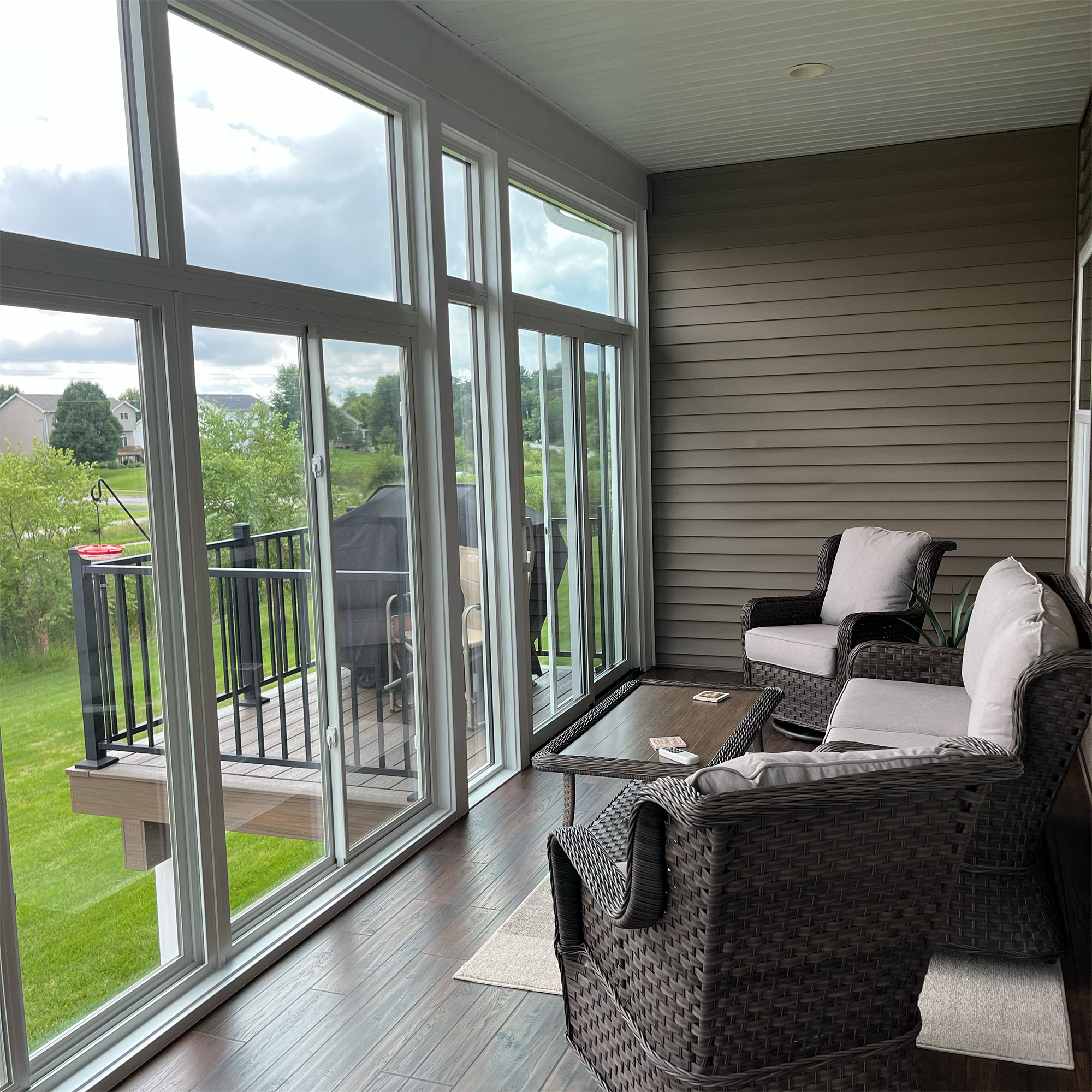 Enclosed sunroom with large windows, wicker furniture with cushions, a coffee table, and a view of a green backyard and deck with barbecue grill.