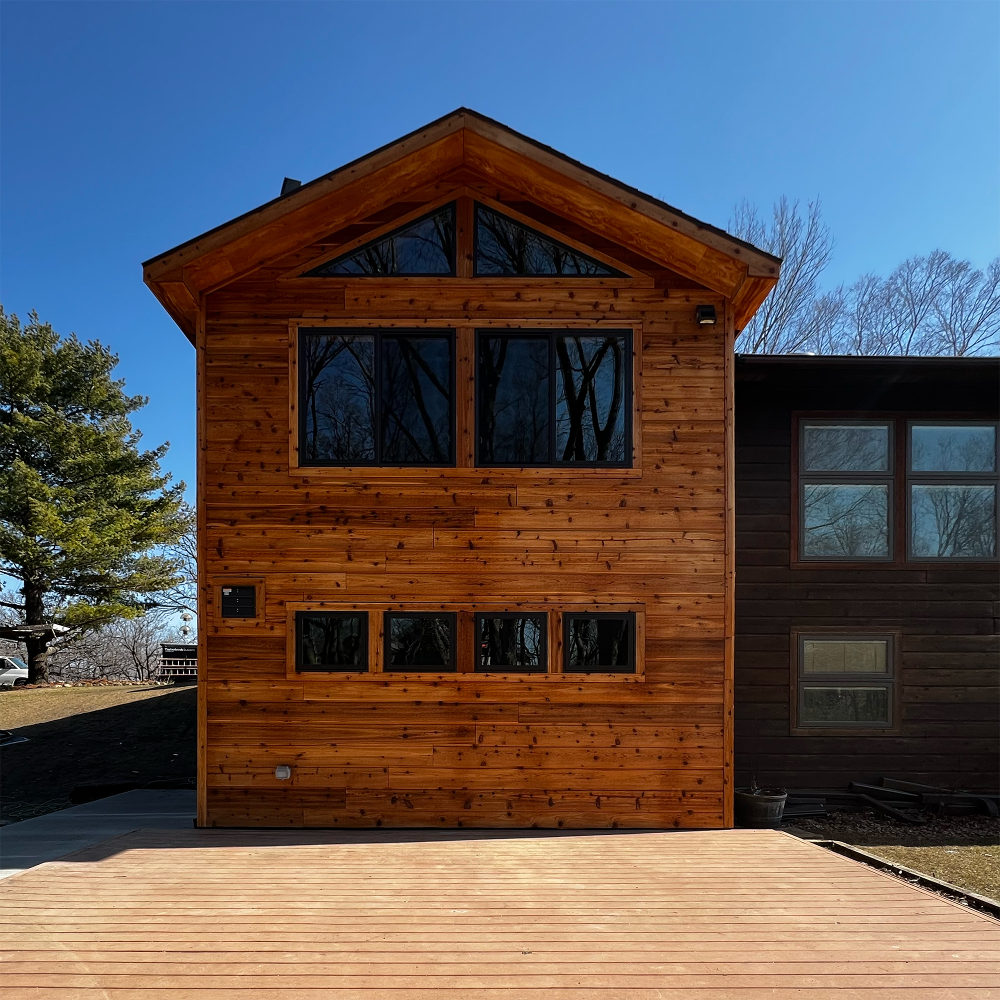 Front view of a wooden cabin addition with large and small windows reflecting bare trees under a clear blue sky.