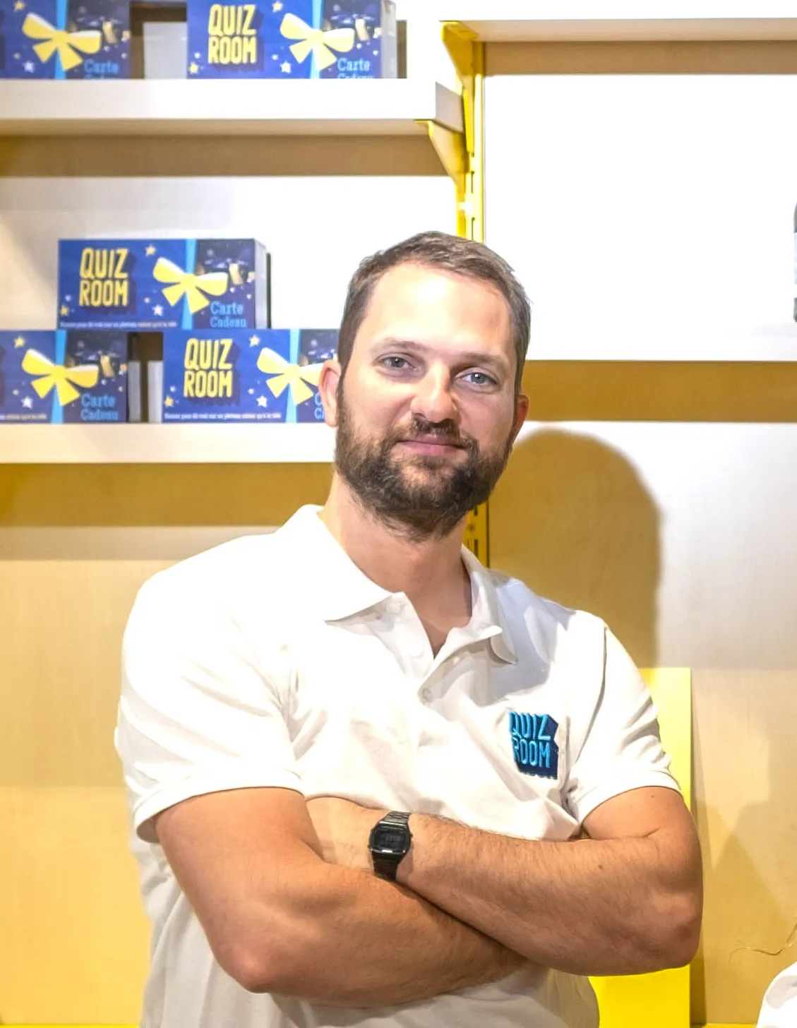 Homme debout avec les bras croisés portant un polo blanc avec un logo Quiz Room, devant des étagères contenant des boîtes Quiz Room.