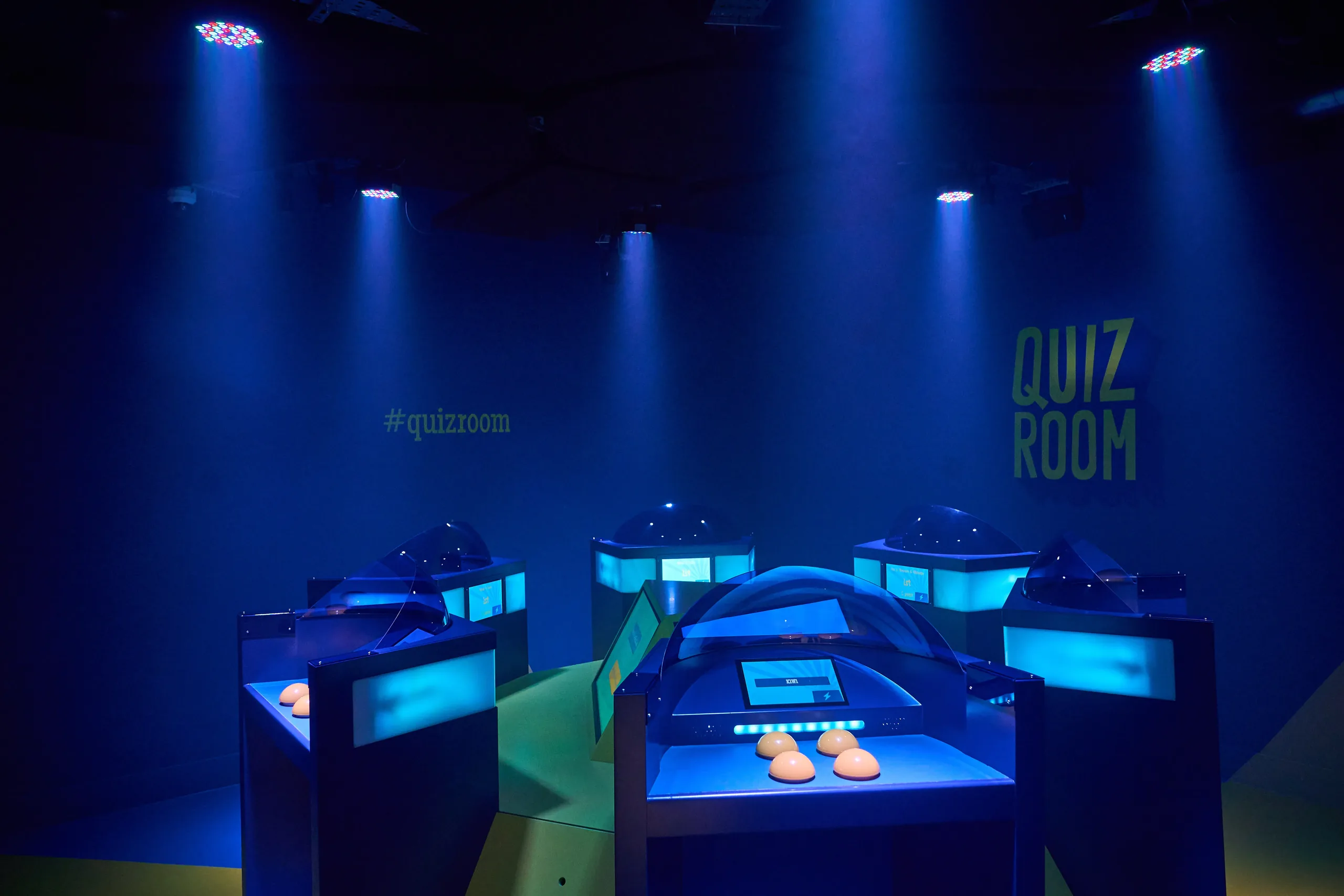 Salle de quiz en lumière bleue avec plusieurs pupitres interactifs équipés d'écrans tactiles et de boutons orange sous des spots lumineux.
