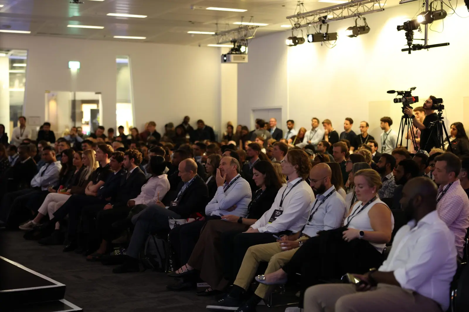 A packed room of attendees watching a session, with camera crew at the back