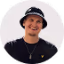 Young man wearing a black hat and black shirt, smiling against a white background.