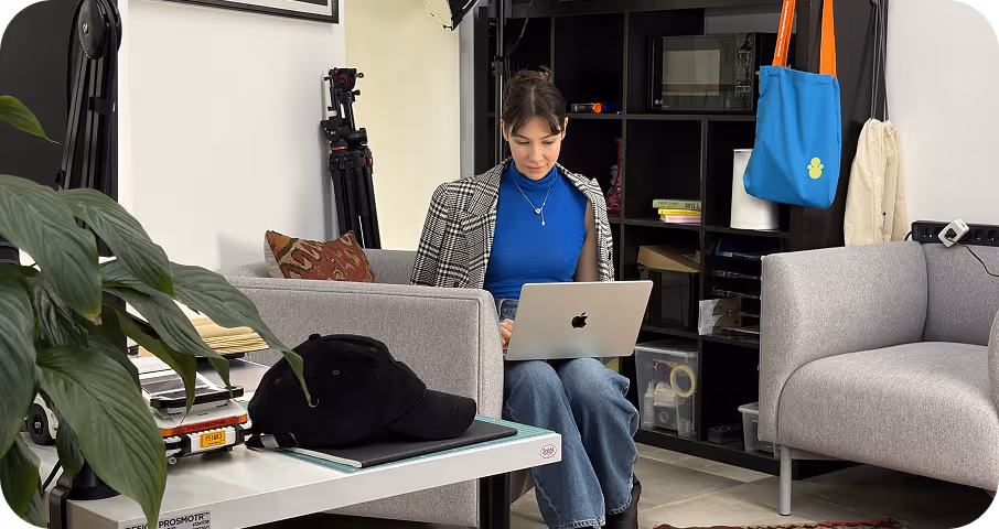 Woman seated on couch working on a laptop in a modern room with shelves and hanging bags.