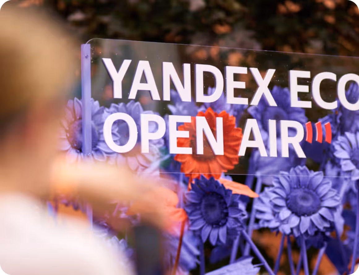 Transparent sign with Yandex Eco Open Air text in front of purple and orange flowers outdoors.