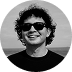 Smiling person wearing sunglasses and a dark shirt against a blurred outdoor background.