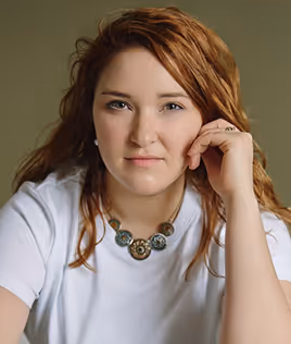 Woman with red hair resting her face on her hand, wearing a white shirt and decorative necklace.